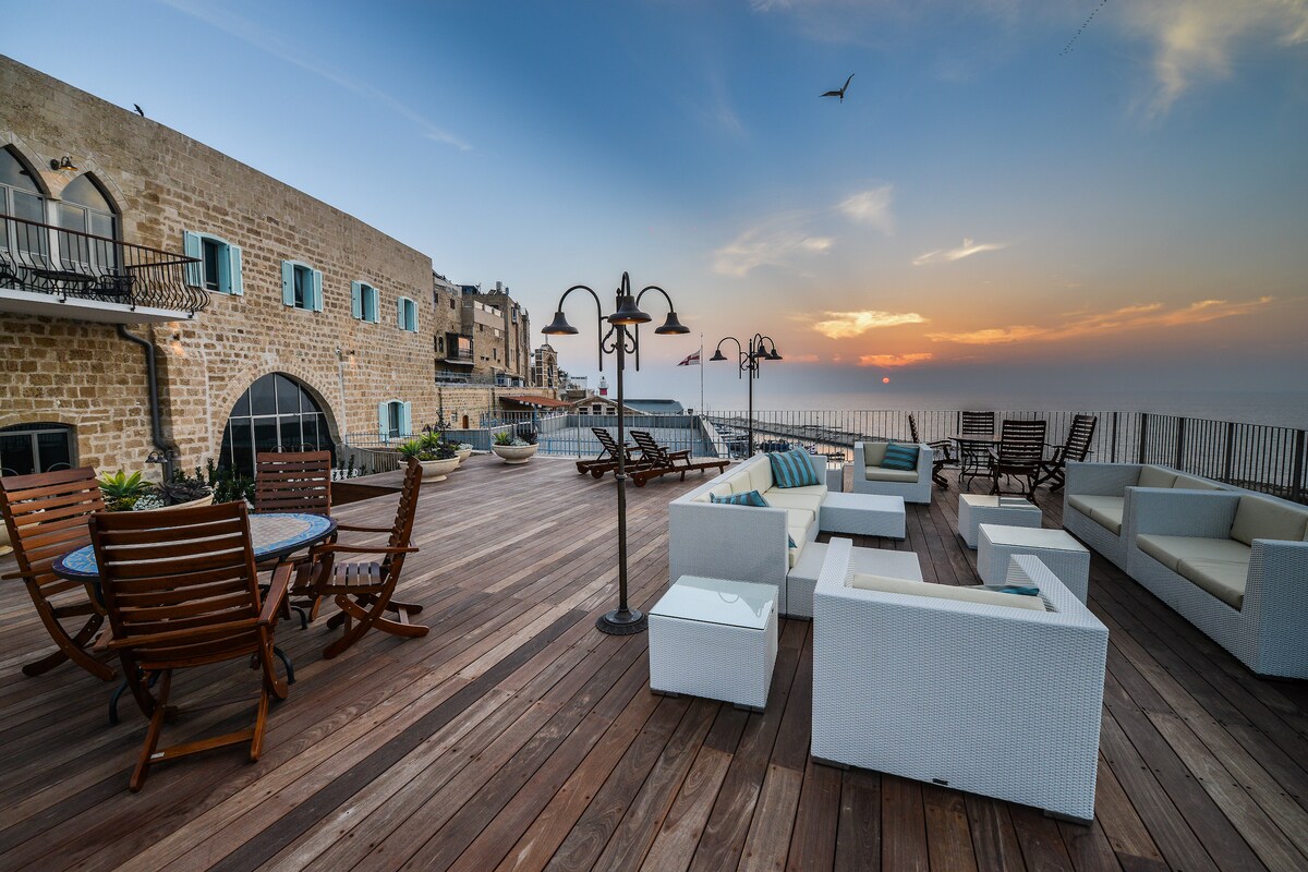 A spacious terrace features wooden flooring, surrounded by elegant seating arrangements. Lounge chairs and sofas invite relaxation, while a dining table with wooden chairs is positioned to take in the views. The sunset casts a warm glow over the scene, enhancing the coastal ambiance.
