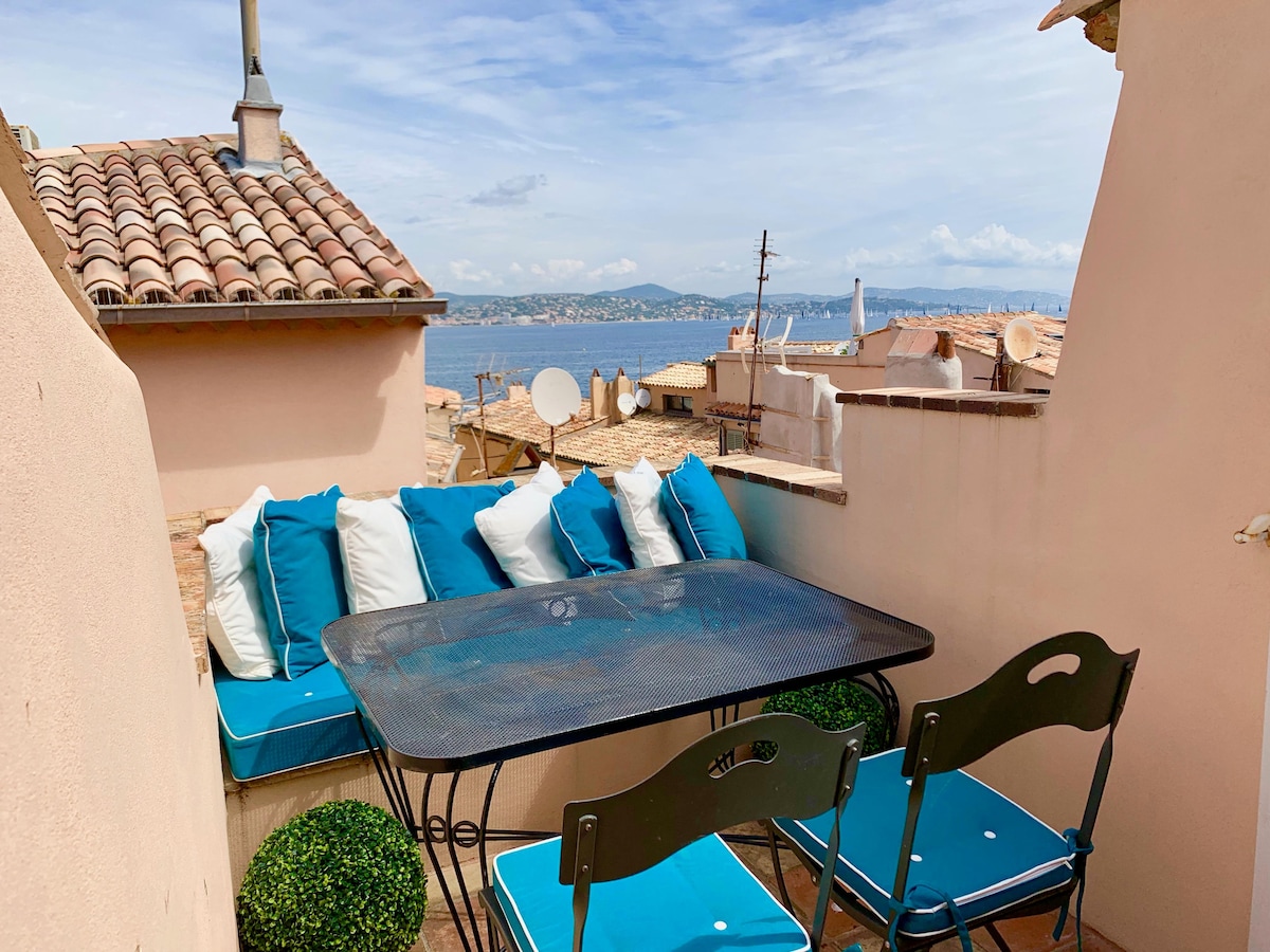 A terrace area features a dining table surrounded by cushioned seating and green potted plants. A scenic view of the sea is presented in the background, with hints of rooftops visible. Soft, cloud-dotted skies create a tranquil atmosphere.