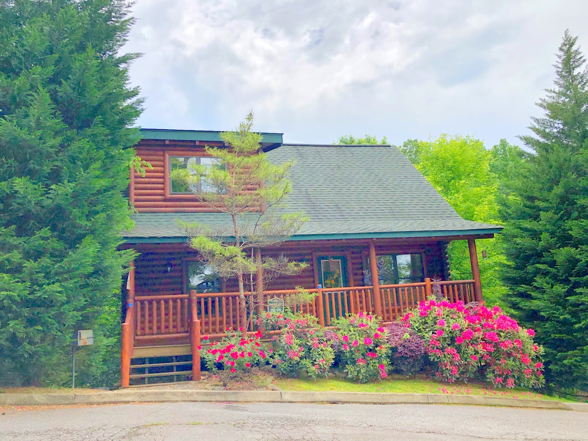 A log cabin is nestled within lush greenery, showcasing a welcoming front porch with wooden railings. Vibrant floral bushes in shades of pink and purple surround the cabin, adding a touch of natural beauty to the setting. A gently sloping driveway leads up to the entrance.