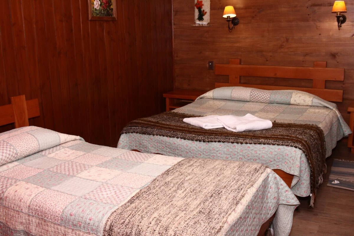 A warm sleeping area features two spacious beds with patchwork quilts and cozy blankets. The wooden walls add to the rustic charm, while wall-mounted sconces provide soft lighting. A neatly folded towel is placed on one of the beds, contributing to a welcoming space.