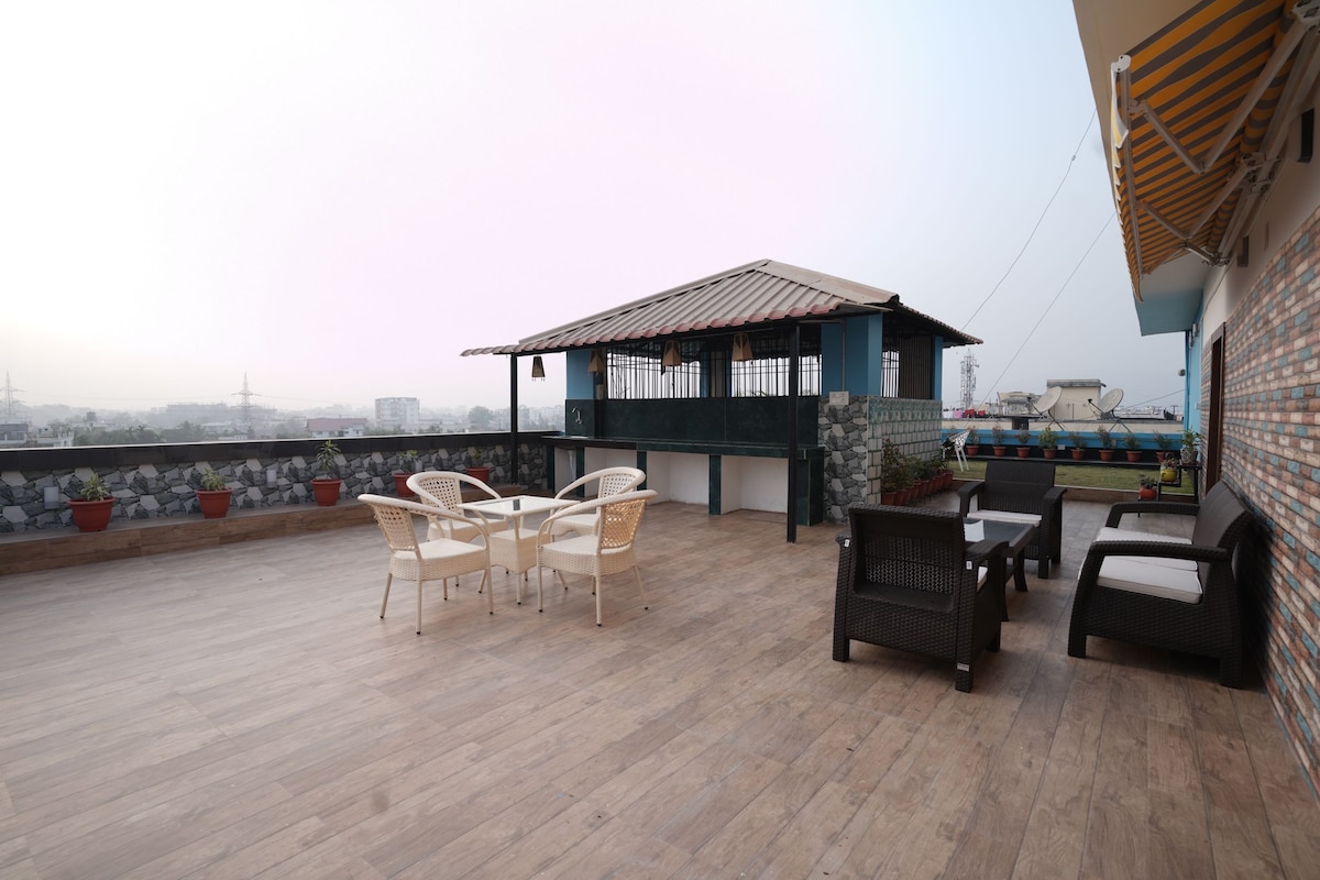 An expansive rooftop terrace features a wooden floor with several outdoor seating arrangements, including a round table with chairs and lounge chairs. A shaded structure provides a gathering space, enhancing the area for family and friends to enjoy the view.