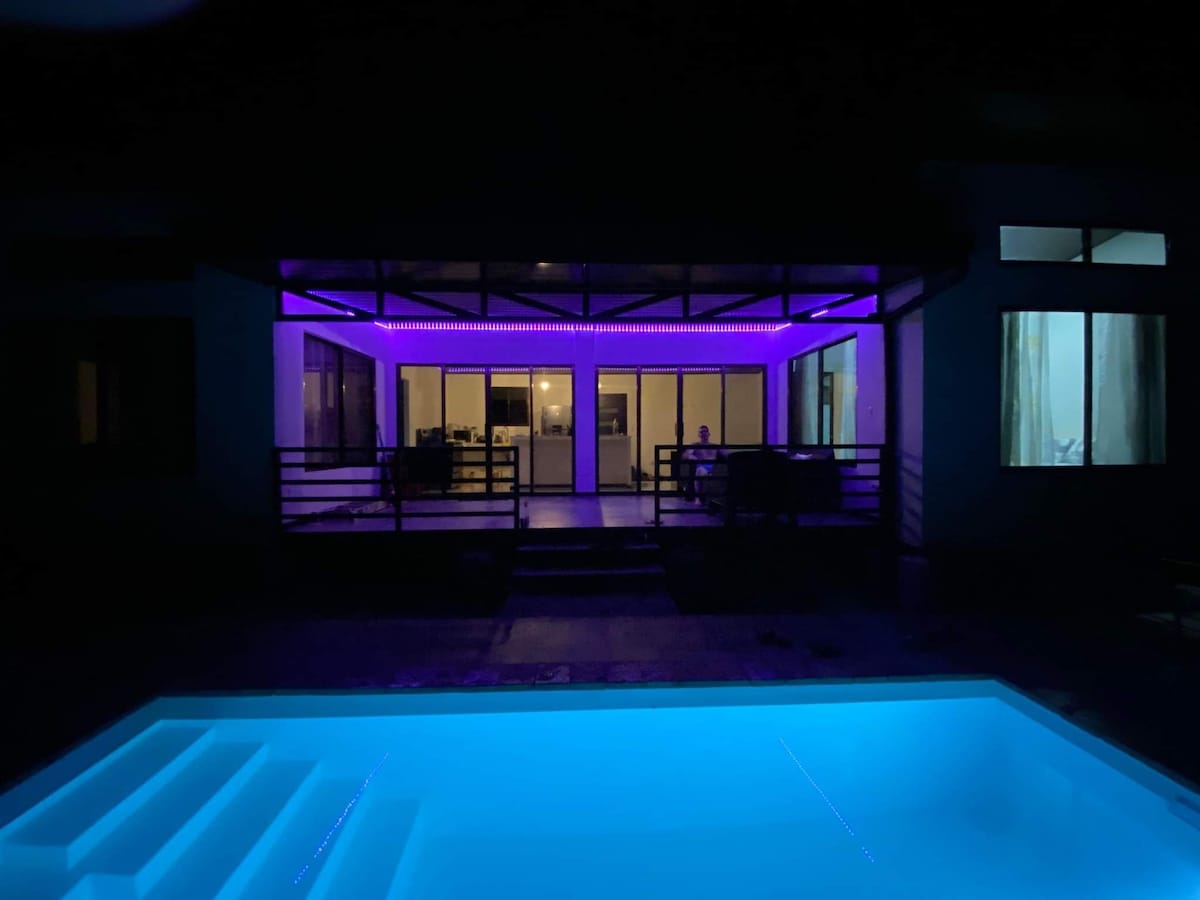 The illuminated pool shines in vibrant blue hues, reflecting the exterior lighting. In the background, large glass doors provide views of the living area, where soft purple accent lights create a welcoming ambiance. Several windows allow glimpses of the interior spaces.