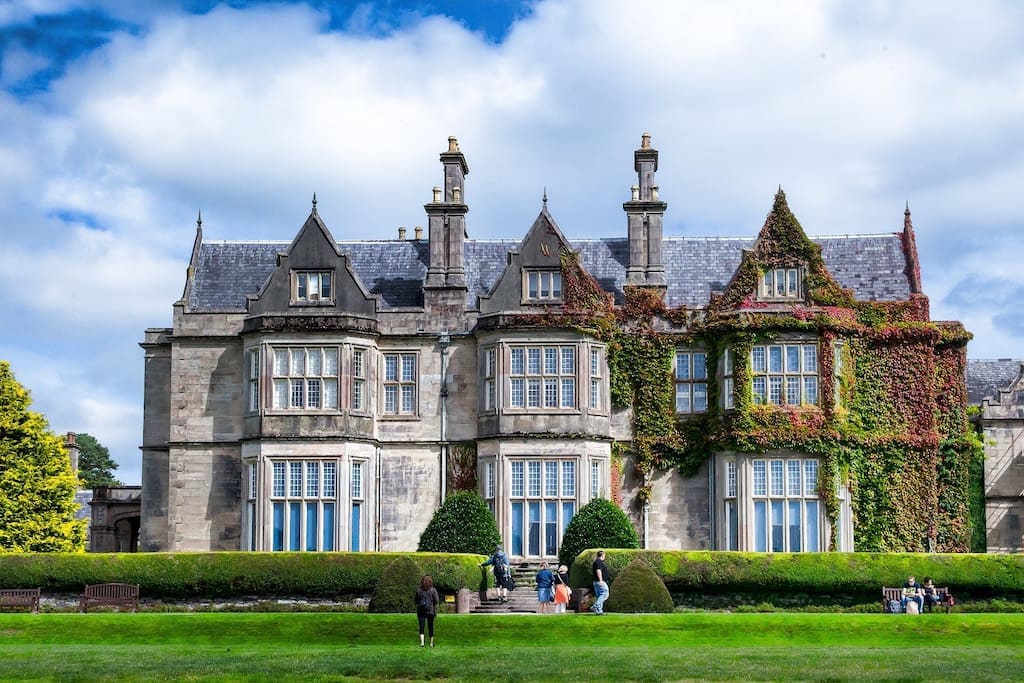 A large historic building showcases architectural features like pointed gables and tall windows. Its façade is adorned with climbing greenery, creating a blend of natural beauty and craftsmanship. A well-maintained lawn with manicured hedges stretches in front, providing space for outdoor gatherings or relaxation.