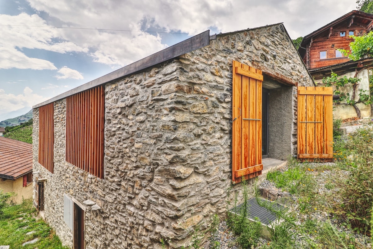 A traditional Swiss mountain barn has been transformed into a modern house, featuring natural stone walls and wooden accents. Large wooden doors on the front open to the surrounding green space, blending the home with the scenic outdoor environment.