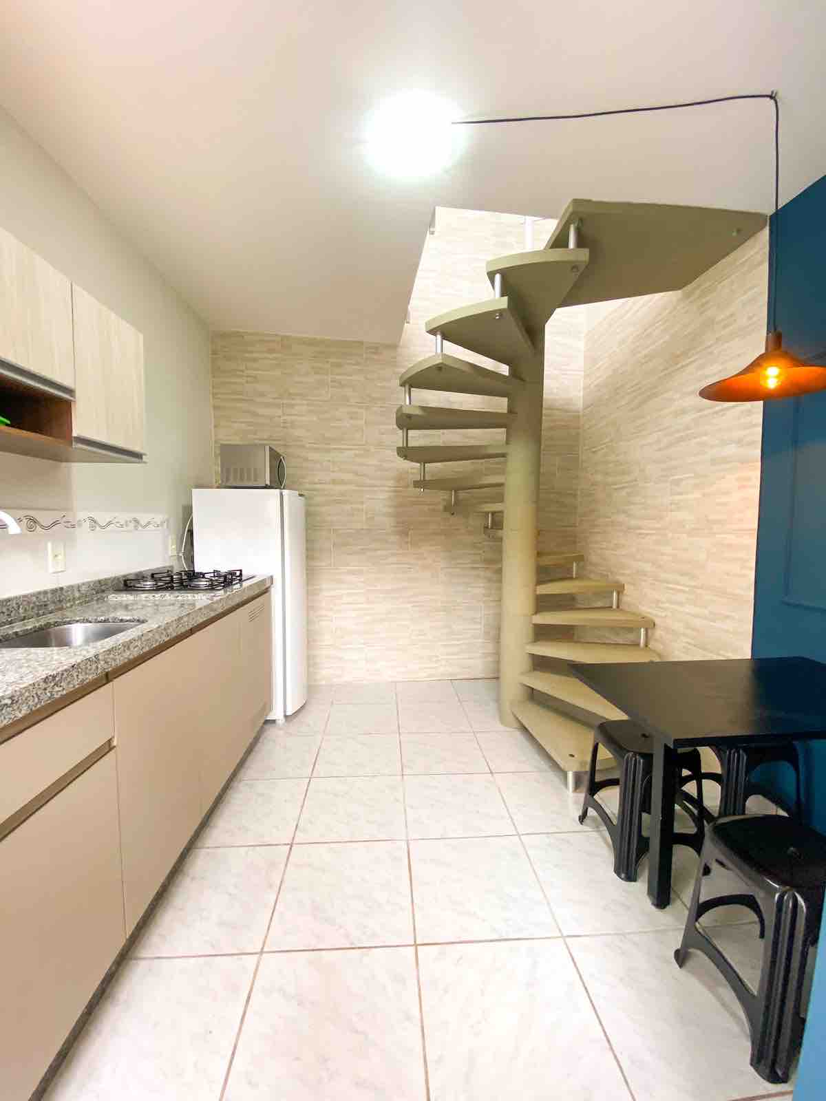 A well-equipped kitchen features light-colored cabinetry and a granite countertop, with essential appliances including a refrigerator and a gas stove. A modern spiral staircase leads to the upper level, while a small dining area is provided with a table and two chairs.