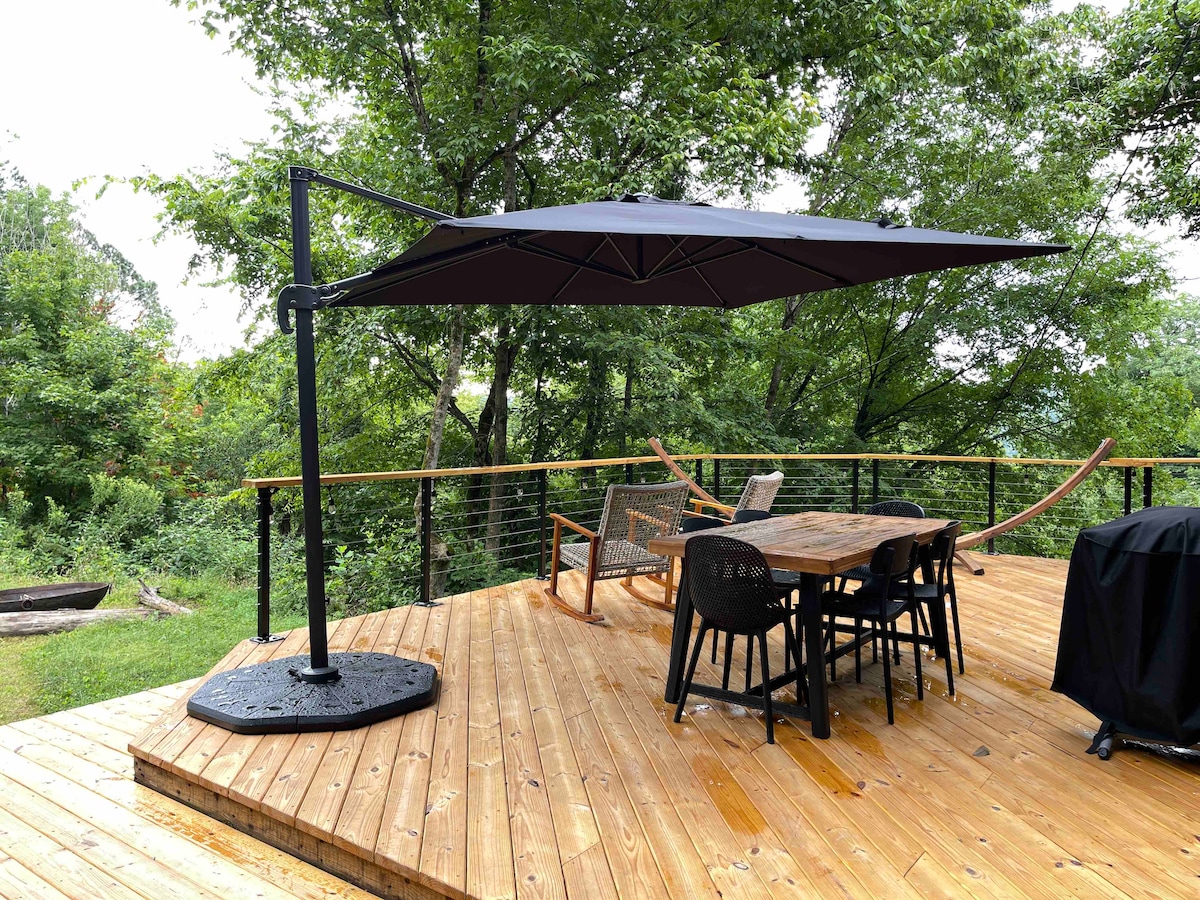 A wooden deck features a large, adjustable umbrella providing shade over a dining table and chairs. Nearby, a grill is positioned for outdoor cooking. The surrounding area is lush with greenery, enhancing the natural setting of the spacious outdoor space.