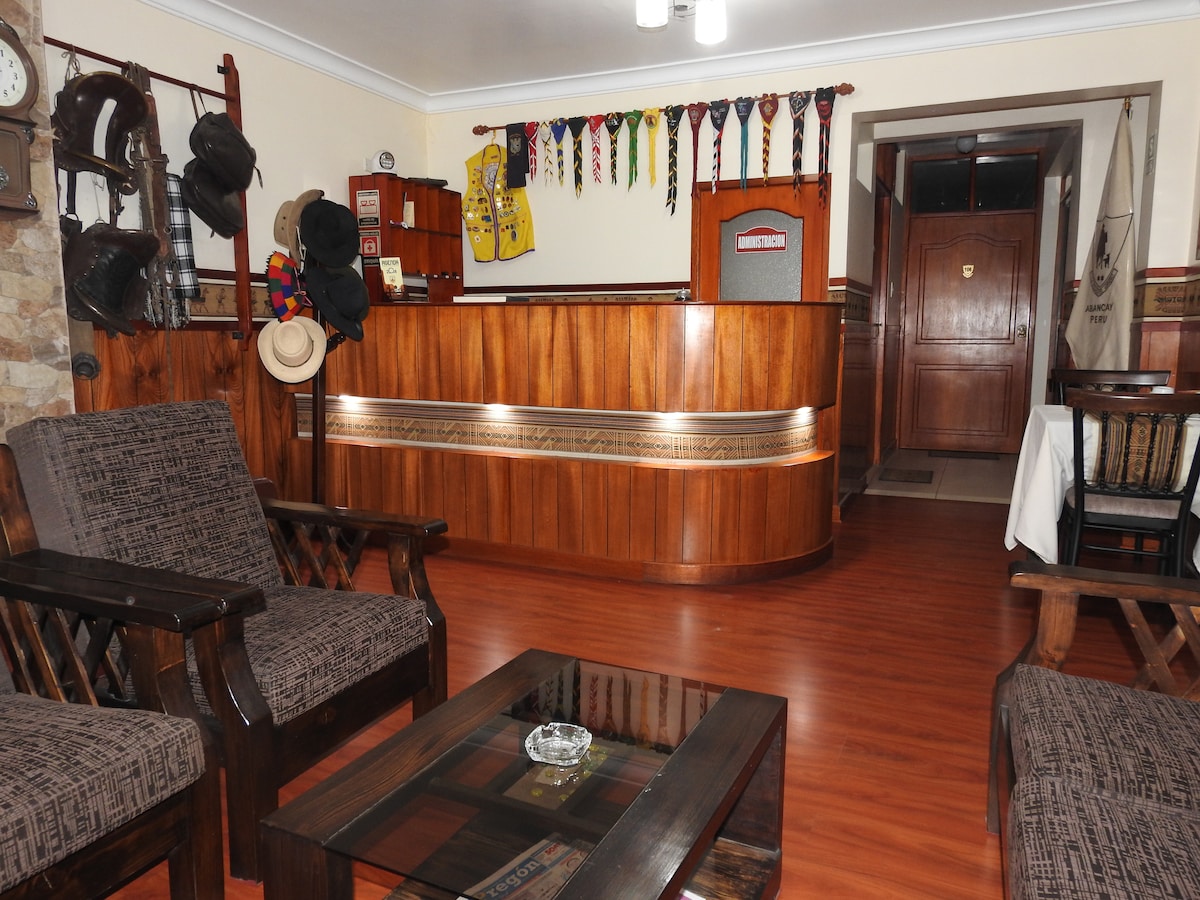 The reception area features a wooden front desk with decorative elements arranged on the wall. Comfortable seating is provided by dark wooden chairs with patterned cushions, complemented by a coffee table. Light wood accents contribute to a welcoming environment.