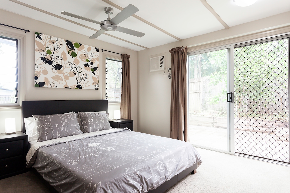 The private bedroom features a queen-size bed with a neutral-colored bedspread. Large windows invite natural light, framed by flowing curtains. A ceiling fan ensures comfort, while a wall-mounted air conditioning unit is positioned for convenience. A decorative wall art piece adds subtle visual interest.