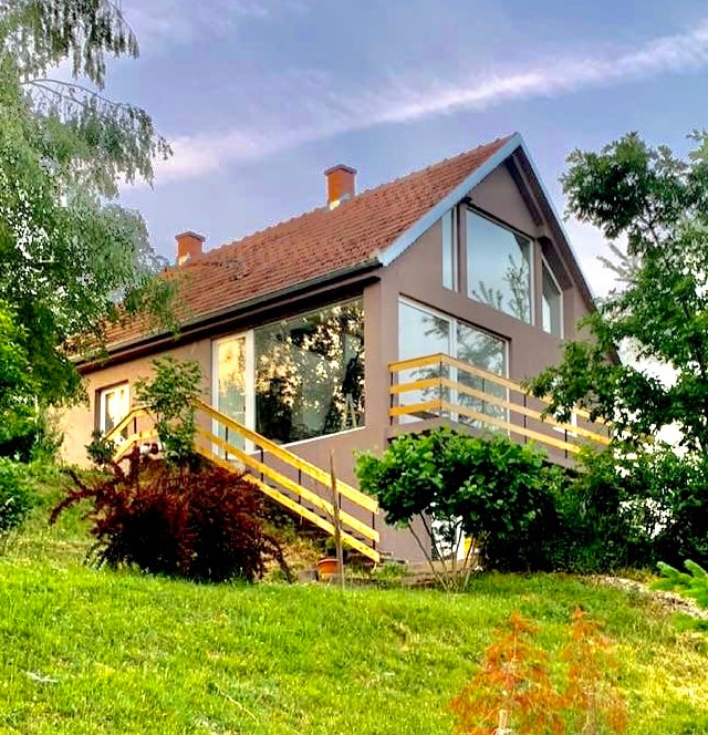 The house is presented with a modern A-frame design, featuring large glass windows that provide clear views of the surrounding greenery. Stairs lead up to a deck with wooden railings, enhancing accessibility to the inviting entryway.