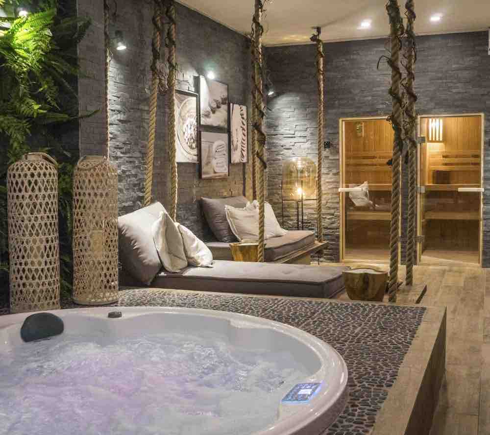 A serene wellness area features a round hot tub beside a wooden seating area adorned with plush cushions. Textured stone walls provide a calming backdrop, while warm lighting creates an inviting atmosphere. A sauna is visible in the background, enhancing the relaxation experience.
