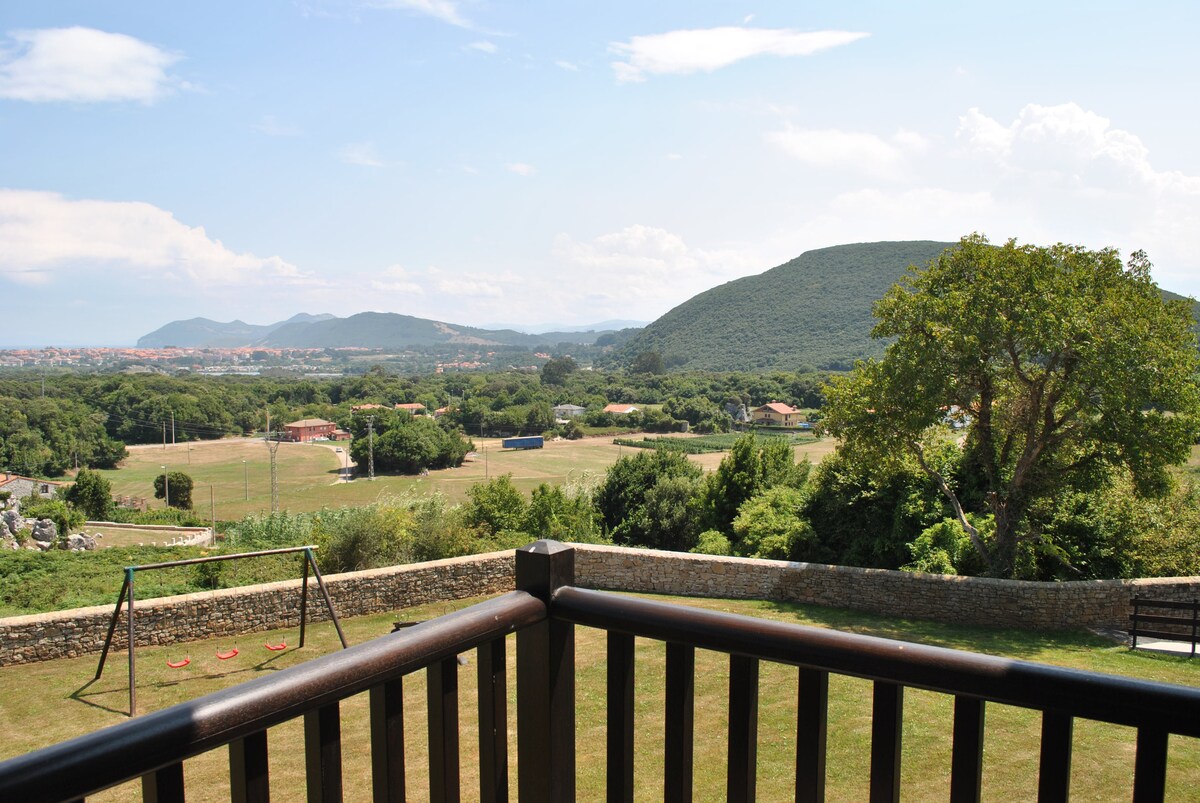 A view from the balcony reveals a expansive landscape featuring rolling hills and greenery. In the foreground, a wooden railing frames the scene, while a playground swing set is visible on the left. The horizon showcases a distant town under a clear blue sky.