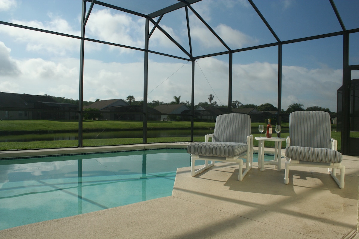 A screened terrace features a private swimming pool with two cushioned lounge chairs positioned beside it. A small table holds two glasses, offering a view of the tranquil water and greenery beyond. The setting provides a quiet space for relaxation.