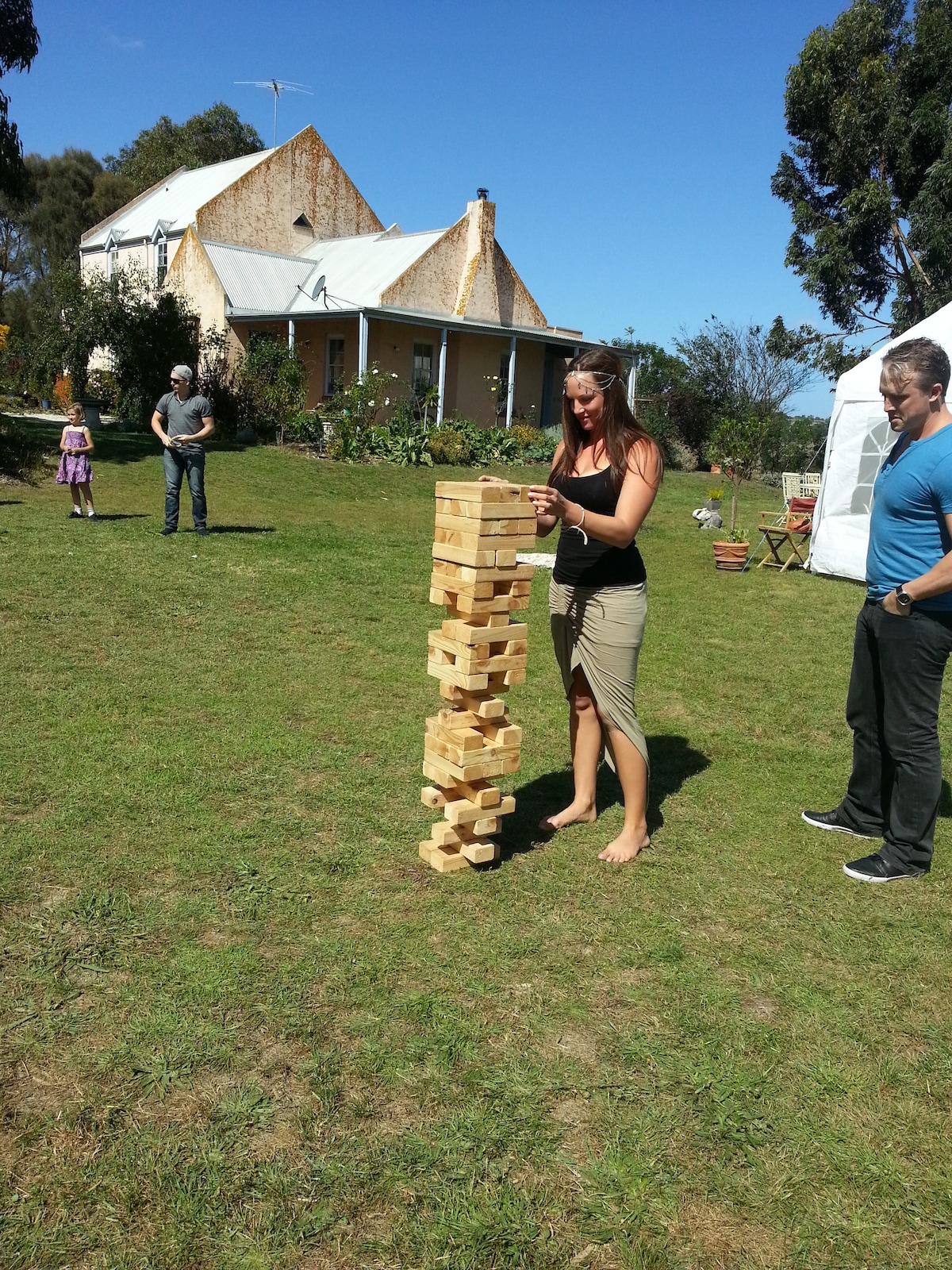A outdoor scene features a person carefully balancing wooden blocks in a game of Jenga. A rustic building is visible in the background, surrounded by greenery and flowering plants. Clear skies and natural light create a relaxing atmosphere.
