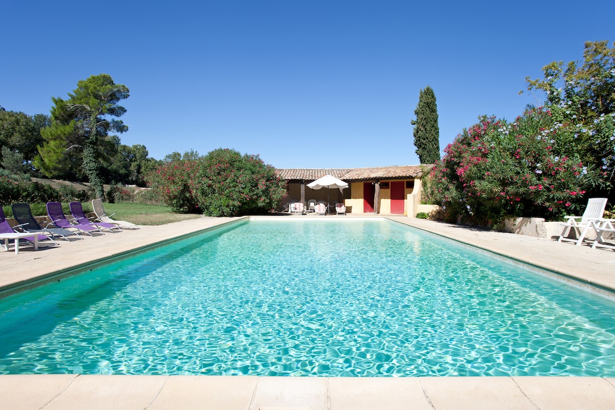 A large swimming pool glistens under a clear blue sky, surrounded by sun loungers and vibrant greenery. The area features a shaded seating space with umbrellas, providing a relaxing spot to enjoy the tranquil environment. Colorful flowering plants add to the serene landscape.