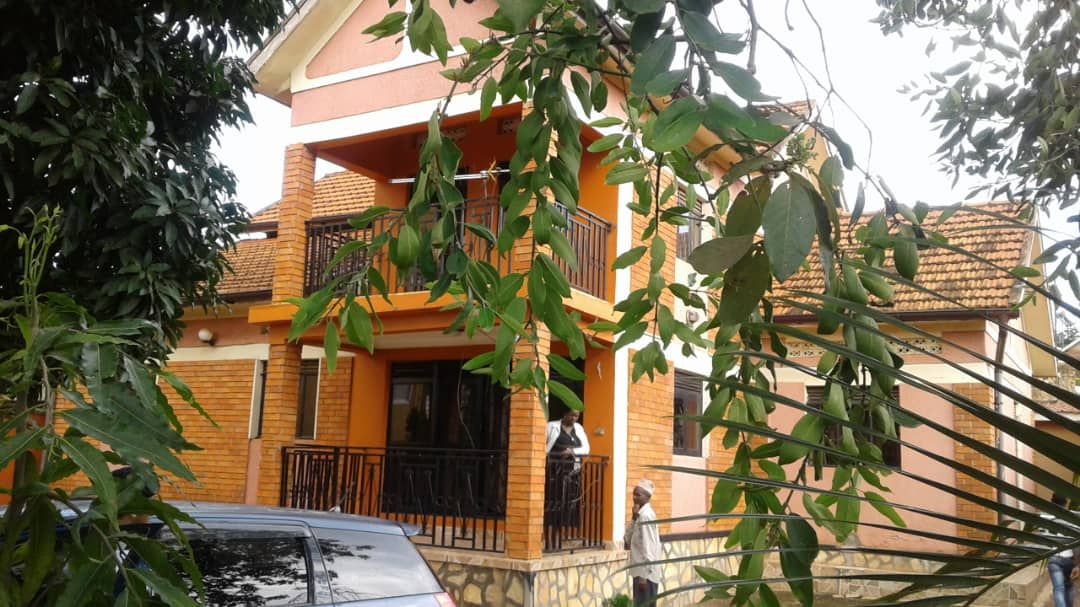 A two-story house displays orange brick exteriors, accented by dark wood balconies. Lush green foliage frames the scene, with a vehicle parked nearby. A person can be seen standing casually on the balcony, while the surrounding trees add a natural touch.