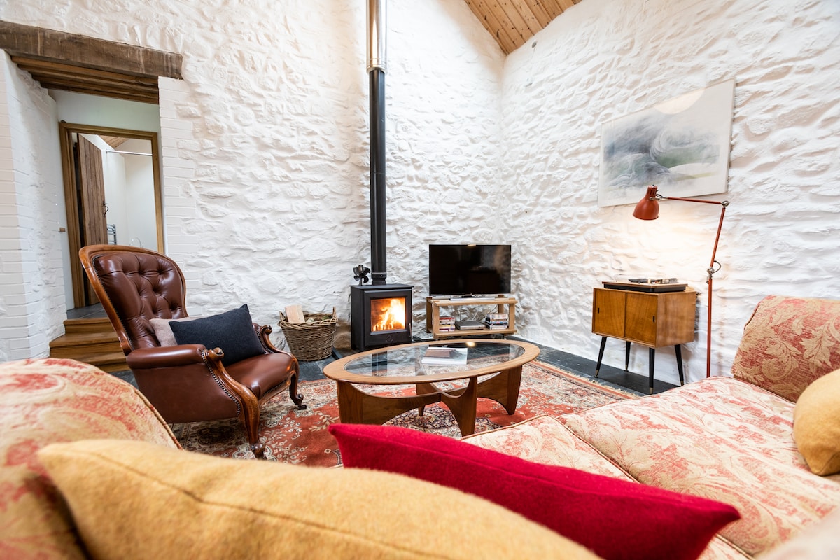 An open living area features textured white stone walls and a wooden ceiling. A brown leather armchair is positioned beside a wood-burning stove, with a small television and record player visible nearby. A large sofa is adorned with colorful cushions, enhancing the cozy ambiance.