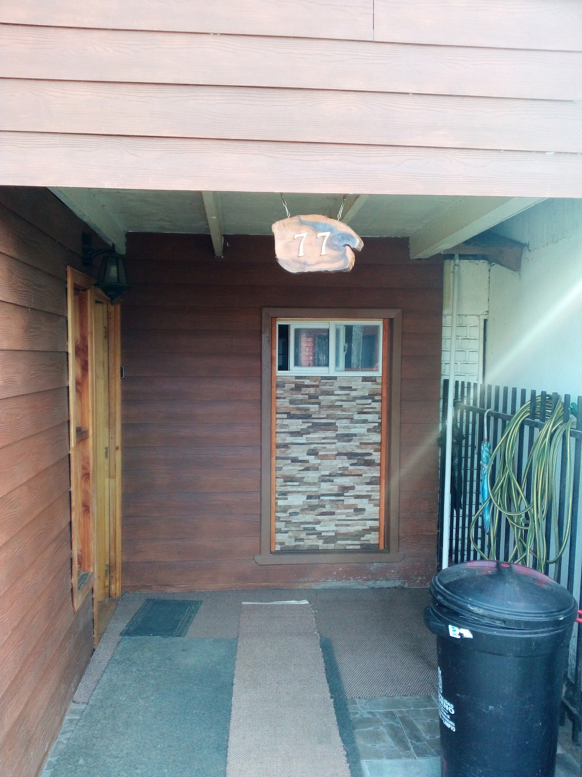 The entrance features a wooden facade complemented by a stone-inlay wall. A house number is displayed prominently above the door, alongside a window that allows natural light. A welcome mat lies underfoot, and a trash bin is positioned nearby.