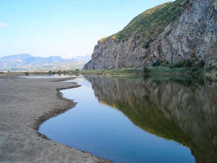A serene lagoon reflects the surrounding hills and cliffs. Gentle sandy shores blend into the calm water, while lush greenery can be seen along the banks. The landscape is enhanced by distant mountains under a clear blue sky.