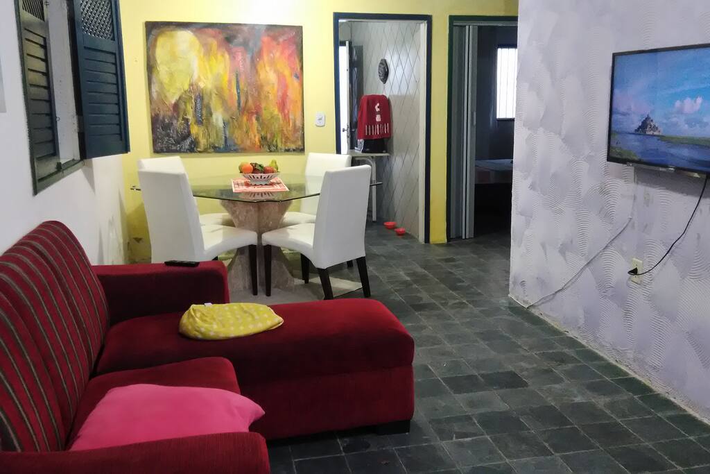sala de estar com sofá de trés lugares, mesa com quatro cadeiras, dois pufes, tv.
