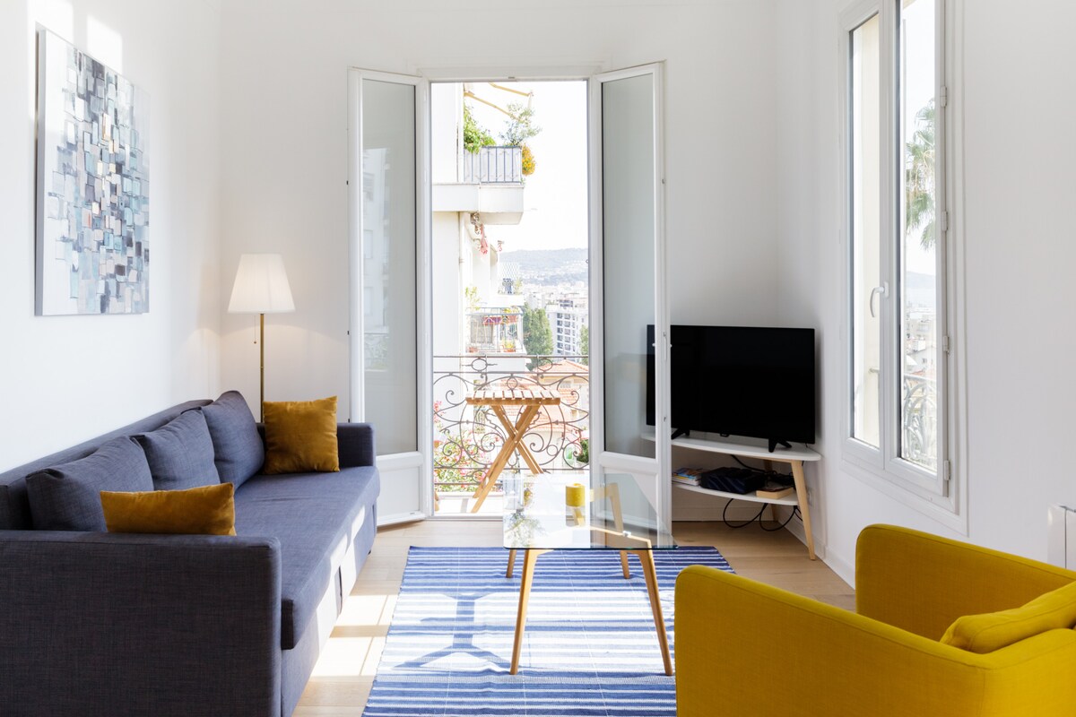 A bright living room features large windows that open to a scenic view. A gray sofa with cushions and a yellow armchair provide comfortable seating. A glass coffee table rests on a striped blue and white rug, while a TV is positioned on a small wooden stand.