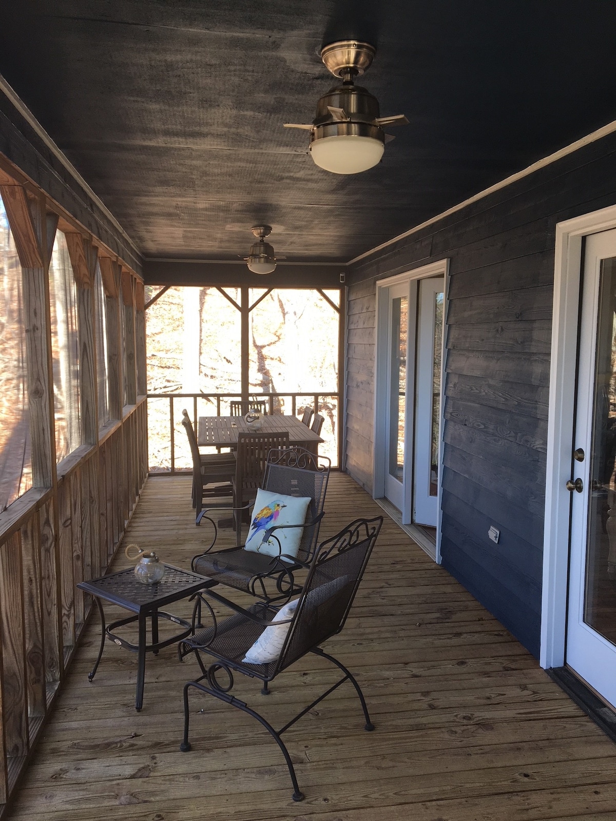 A screened-in porch is shown with wooden flooring and dark blue walls. Several metal chairs surround a small table, creating a comfortable seating area. Natural light filters through the screens, highlighting a peaceful view of the outdoors.