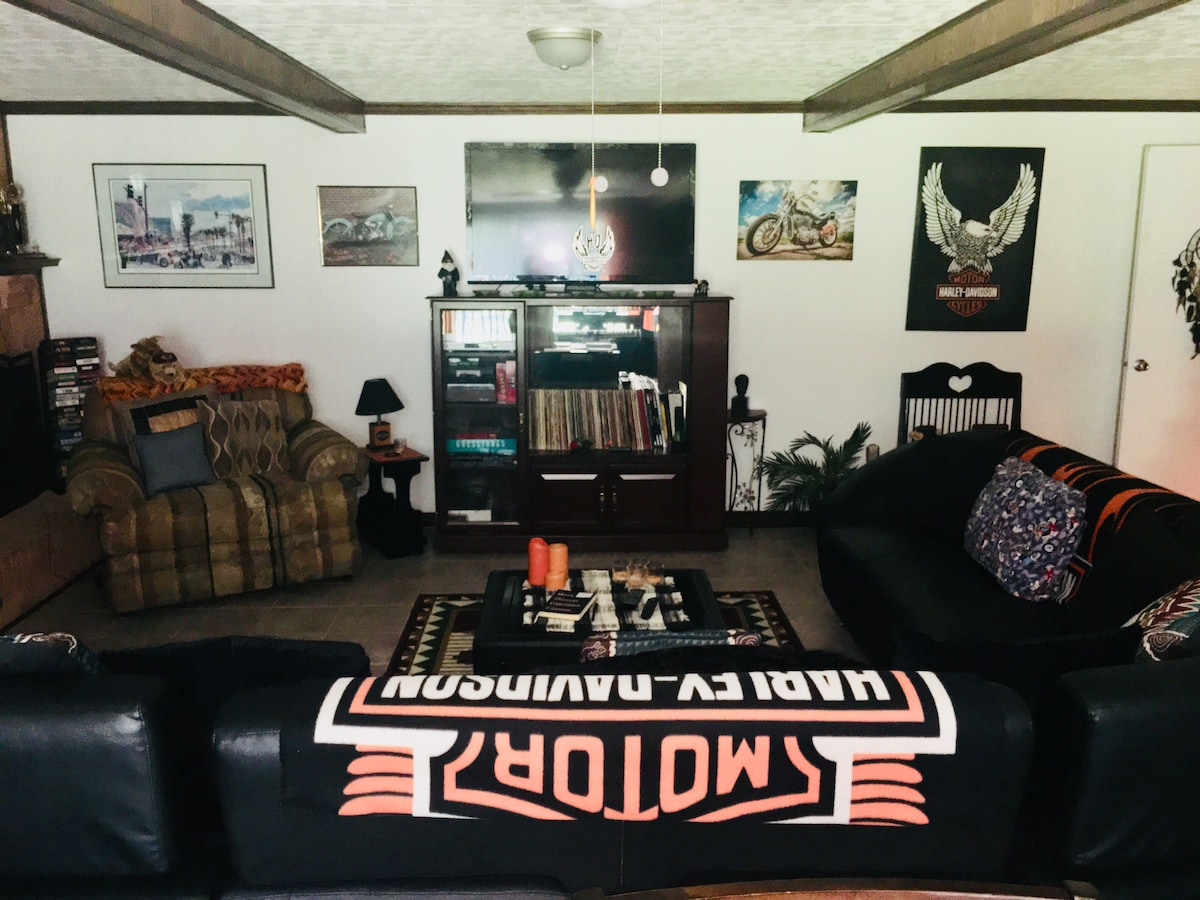 The living room showcases a comfortable arrangement of furniture, including a patterned sofa and armchair. A dark coffee table sits in the center, surrounded by decorative elements like artwork and a collection of books on shelves. Warm lighting enhances the inviting ambiance.