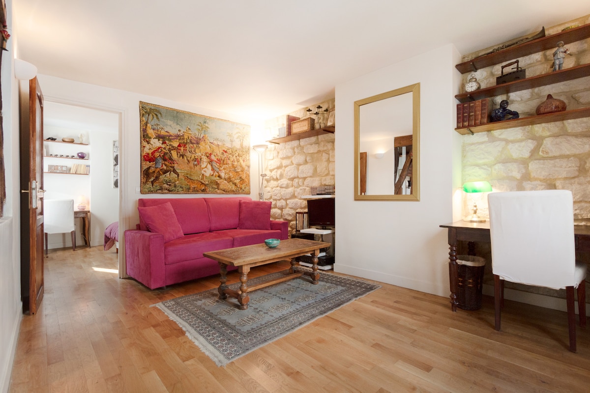A living area is presented with wooden flooring, featuring a vibrant pink sofa and a rustic wooden coffee table. Stone walls add texture, while a large mirror reflects light. A small writing desk with a chair is positioned by a window, enhancing the room's functionality.