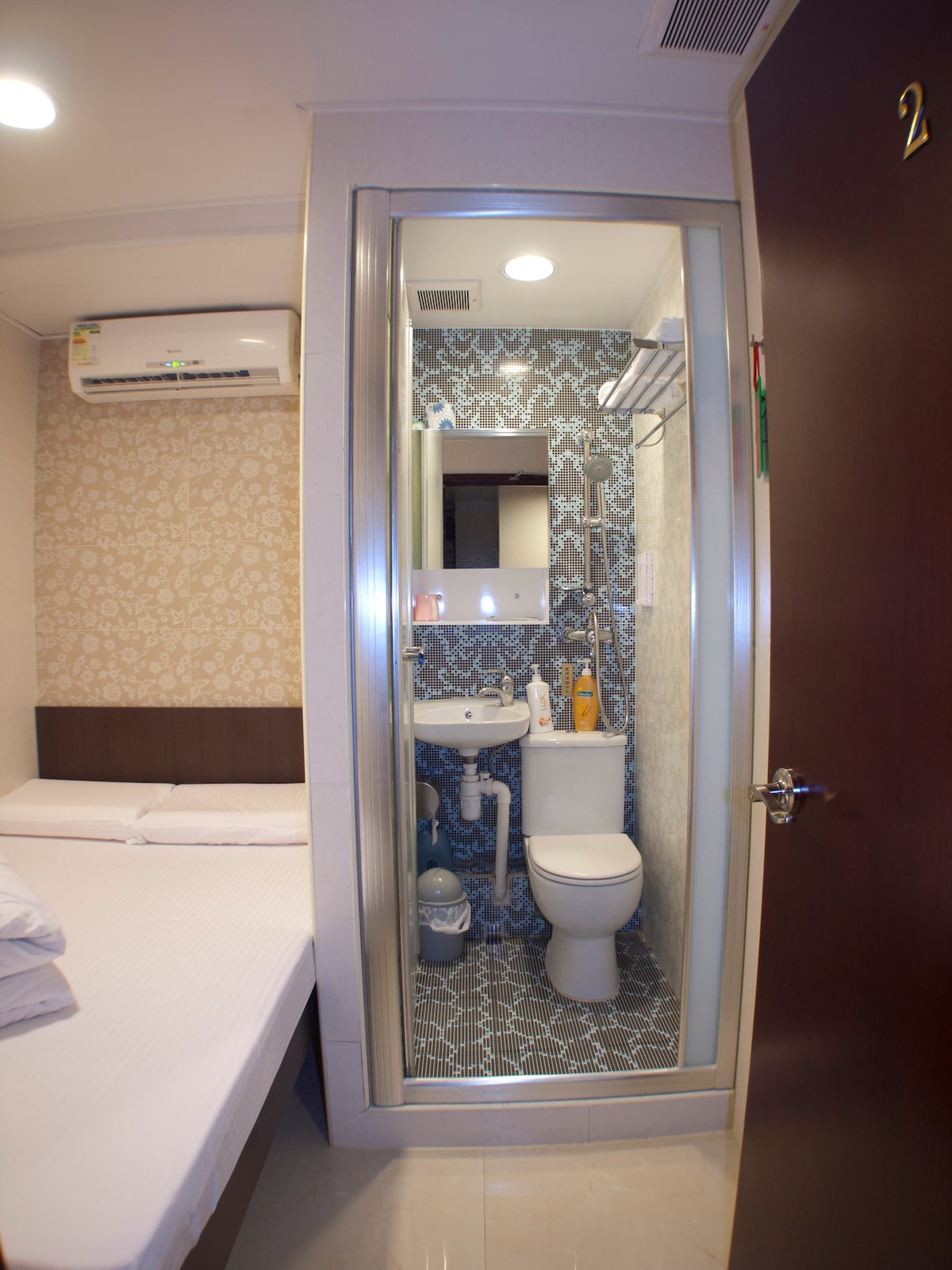 A compact double room features a spacious bed and a private bathroom. The bathroom, visible through a glass partition, includes a shower, sink, and toilet. Wall designs and lighting create a clean, modern appearance. An air conditioning unit is installed above the bed.