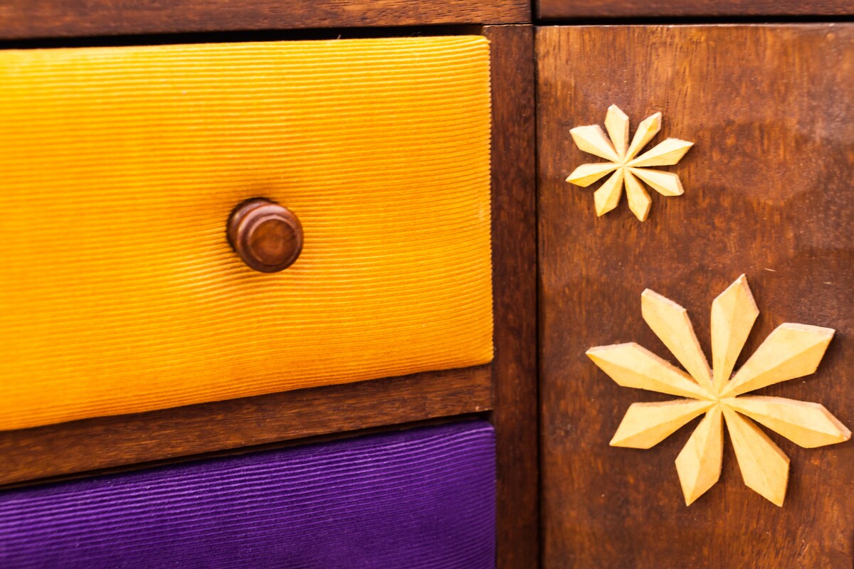 A close-up of a wooden dresser drawer showcases intricate craftsmanship. The deep purple and vibrant yellow drawers feature texture variations, and decorative floral carvings in a lighter wood color add a unique touch. A round wooden knob provides functionality.