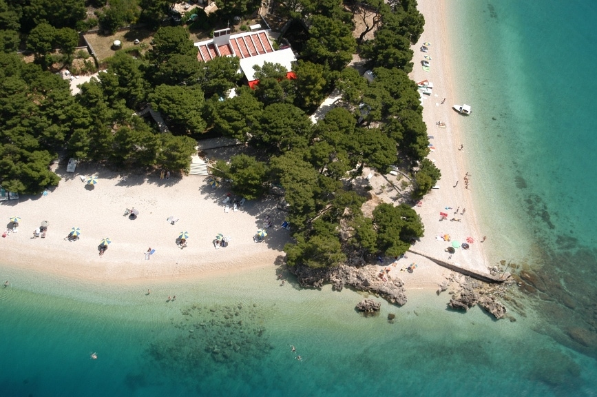 An aerial view captures a sandy beach bordered by a lush pine tree forest. Sun loungers and umbrellas dot the shore, while calm, clear waters gently lap at the sand. The scene includes boats moored in the distance, blending harmoniously with the natural surroundings.