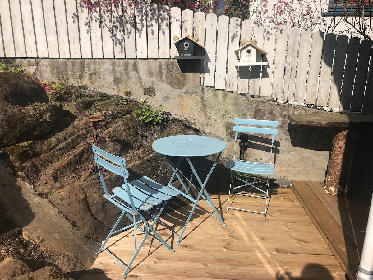 A small outdoor area features a light blue bistro table and two matching chairs, positioned on a wooden deck among natural rock formations. Two birdhouses are visible mounted on the wooden fence behind, surrounded by greenery and colorful plants.
