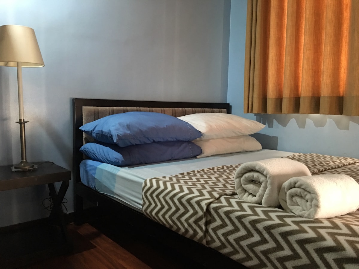 A well-arranged sleeping area features a spacious bed with layered bedding in blue and white. Two neatly rolled towels are placed at the foot of the bed. A bedside lamp and a brown chevron-patterned blanket add subtle comfort, while bright curtains soften the natural light.