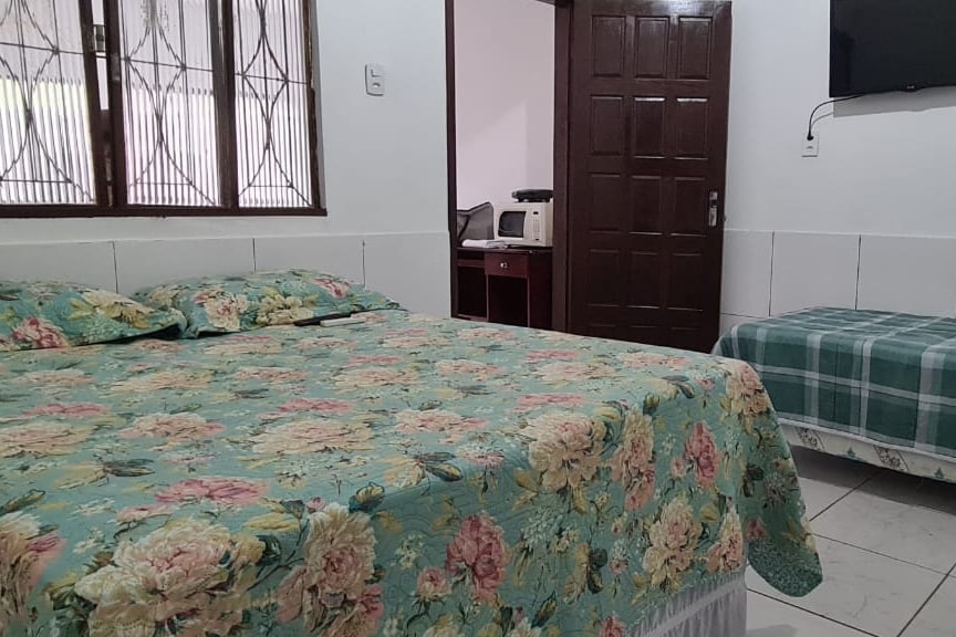 A cozy sleeping area features a spacious bed adorned with a floral patterned cover. A single bed is positioned nearby, while a small kitchenette with a microwave is visible in the background. Natural light enters through the window, which is fitted with decorative bars.