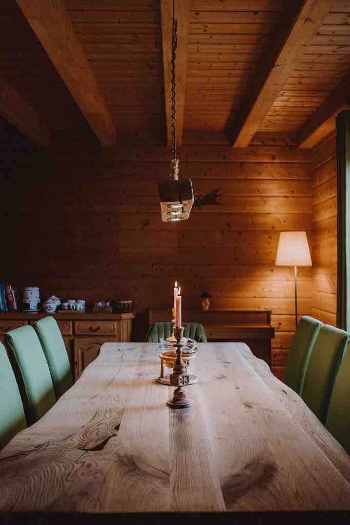 A wooden dining table is centrally positioned, accompanied by several green chairs. Three candles are elegantly arranged in the center, providing a subtle illumination. Wooden beams accentuate the ceiling, while a floor lamp stands softly illuminating the corner of the room.