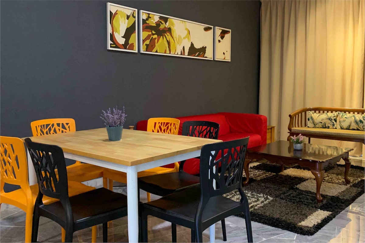 A modern dining area features a wooden table surrounded by six colorful chairs, two in black and four in orange. A red couch is situated nearby, complemented by a traditional rattan chair. Wall art is displayed above the table against a dark grey backdrop.