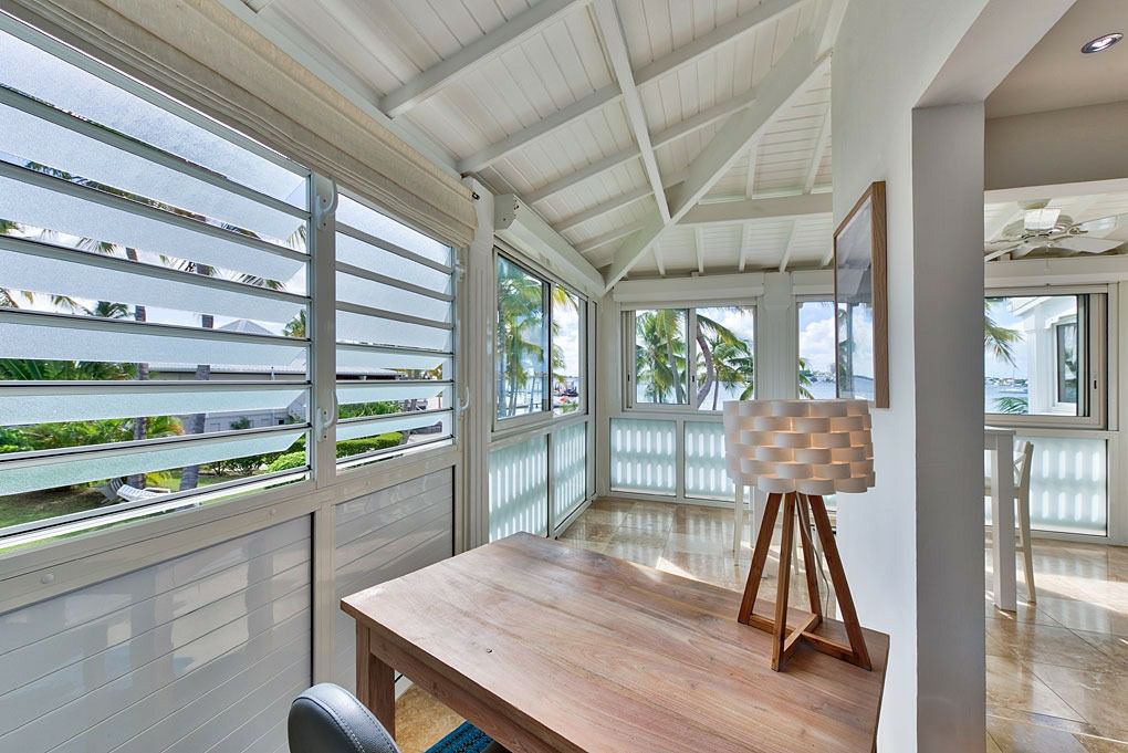 A bright, airy workspace is featured, with large windows that provide natural light and a view of the lush surroundings. The wooden desk is paired with a modern chair, while a decorative lamp adds a touch of style. Palm trees are visible outside.
