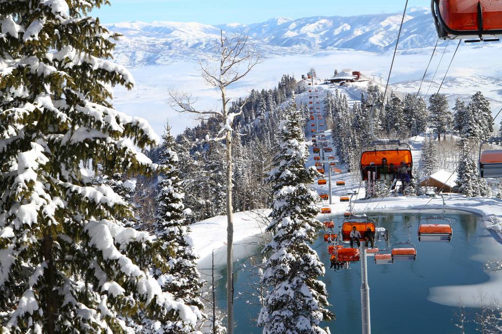A ski lift is visible ascending a snow-covered mountain, surrounded by evergreen trees. The landscape features a mix of white snow and patches of blue sky, with distant peaks creating a serene backdrop.