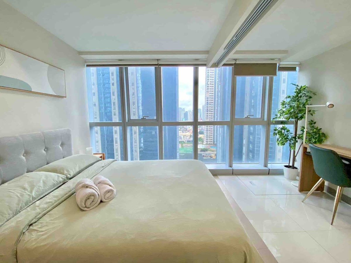 The bedroom features a spacious bed dressed in soft linens, complemented by neatly arranged towels. Floor-to-ceiling windows provide natural light and views of the surrounding buildings. A small desk with a lamp is positioned next to a decorative plant, creating a cozy workspace.