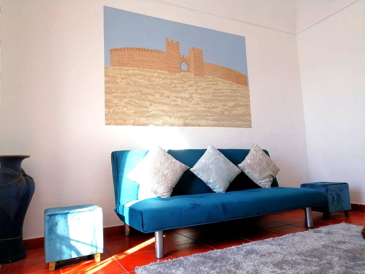 A comfortable seating area is highlighted by a teal sofa adorned with decorative pillows. Blue stools are positioned nearby. A large wall art piece depicting a castle is featured above the sofa, while natural light filters into the space, creating a bright atmosphere.