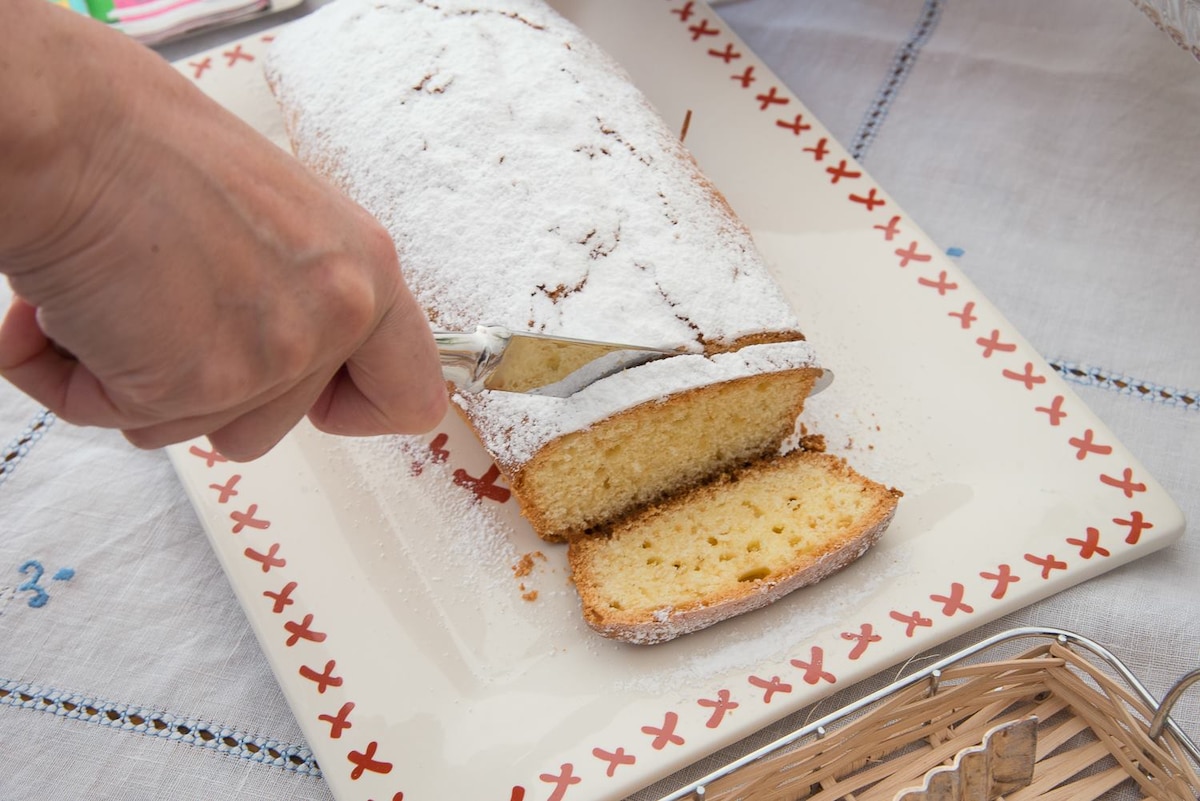 A hand is seen slicing a cake dusted with powdered sugar, resting on a decorative plate with a red border. A soft, sponge-like texture is visible, with a light golden color. The setting includes a patterned tablecloth that adds to the inviting atmosphere.