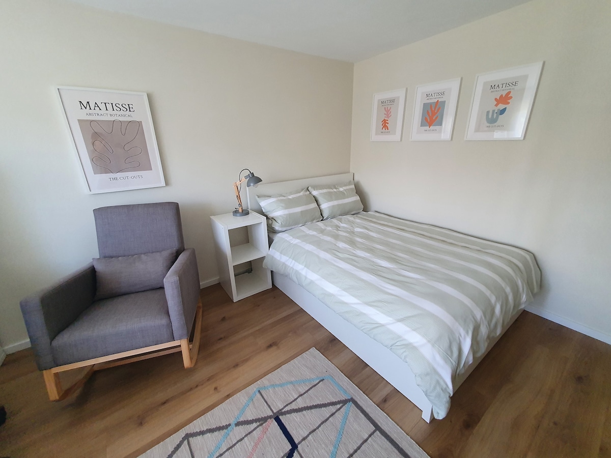 A modern sleeping area features a spacious bed with light-colored bedding. A gray armchair sits beside a white nightstand. Art prints adorn the walls, adding a touch of color to the neutral decor. Light wood flooring enhances the room's warm ambiance.