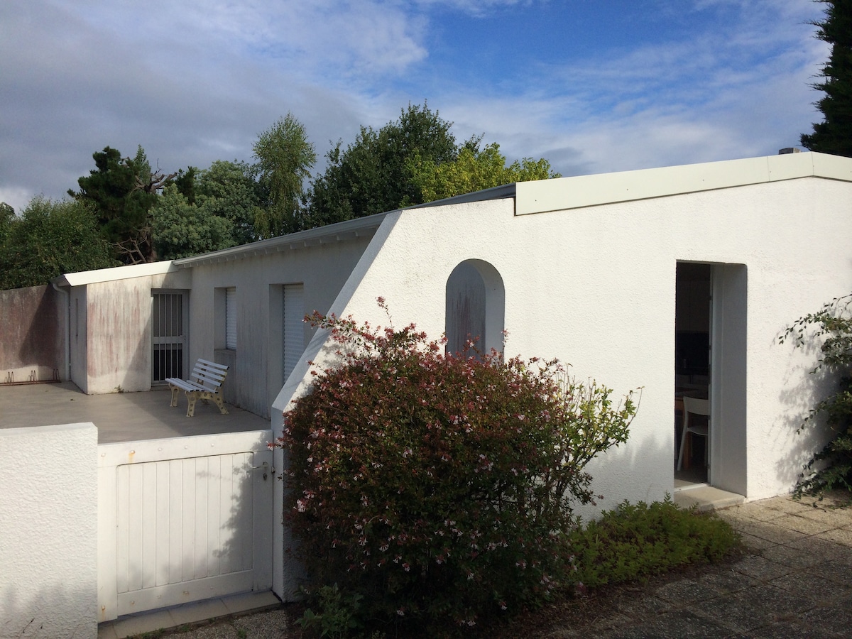 The exterior of the independent house features a clean white façade with a sloped roof. A terrace is situated in front, complemented by green shrubbery and a seating area. Sturdy doors lead into the house, and the surrounding garden adds to the peaceful ambiance.