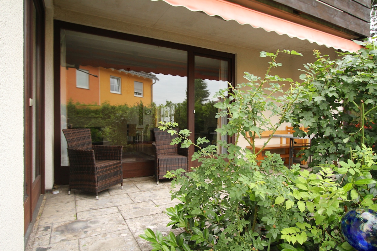 A private terrace features potted greenery and stone tiles. Two wicker chairs are positioned around a small table, offering a cozy seating area. Light filters through the glass doors, which open to the living space, creating a connection between indoor and outdoor areas.