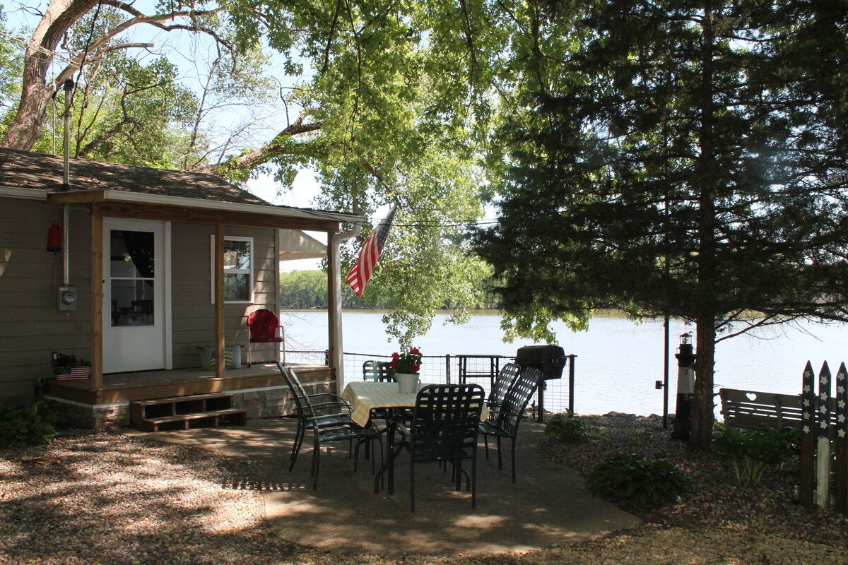A charming cottage exterior is viewed from a shaded patio, furnished with a table and chairs. A flag gently waves in the breeze. Nearby, the calm waters of the Mississippi River create a serene backdrop, framed by lush trees.