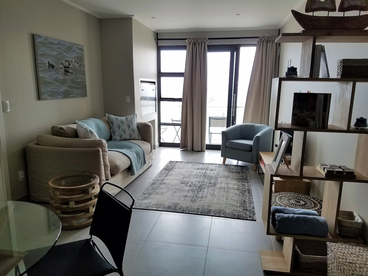 A modern living area features a comfortable sofa draped with a light blanket and a blue accent chair, complemented by a circular table and black chairs. Large glass windows allow natural light, while a textured area rug adds softness underfoot. A decorative shelf displays various items.