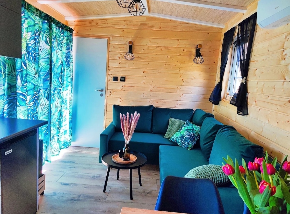 A cozy living area features a dark green sectional sofa adorned with decorative pillows. A round table sits in front of the sofa, complemented by a vase of flowers. Large windows are dressed with tropical-patterned curtains, allowing natural light to brighten the wooden walls.