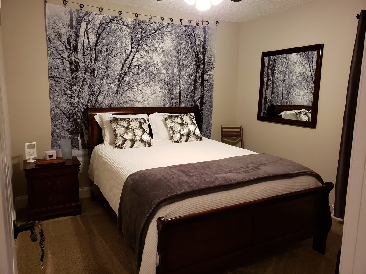 A bedroom features a spacious bed, dressed in white linens with patterned pillows. A large wall mural of winter trees enhances the decor. A wooden nightstand and a chair are positioned near the bed, while a mirror reflects the room's calming ambiance.