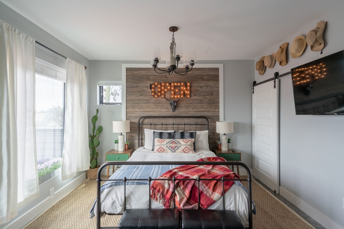An inviting bedroom features a spacious bed adorned with a patterned blanket. A rustic wooden accent wall with an illuminated 'OPEN' sign adds character. Two bedside tables with lamps flank the bed. Natural light filters through sheer curtains, enhancing the warm atmosphere.