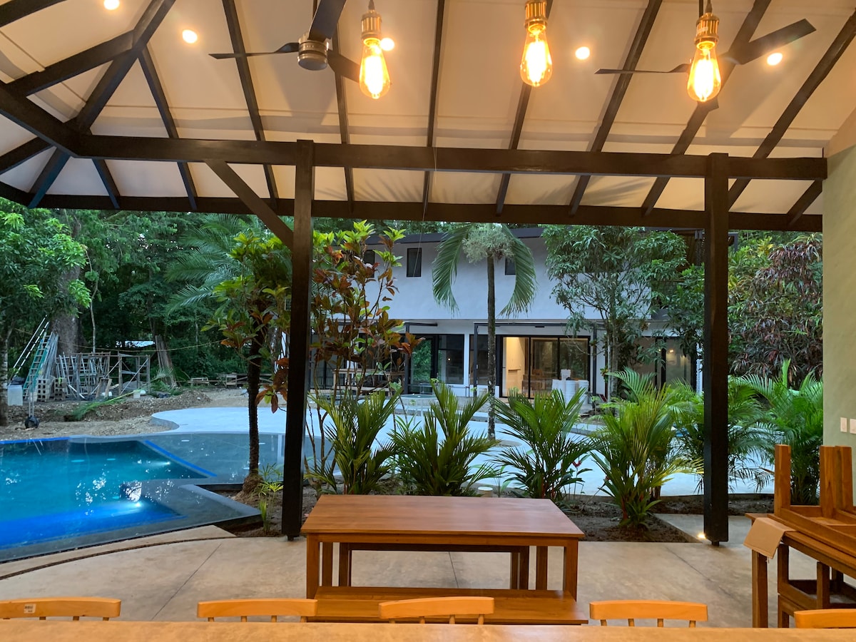 A spacious outdoor dining area is shaded under a roof with exposed beams. A large wooden table is positioned centrally, facing a serene pool surrounded by tropical foliage. The lush greenery and distant view of the property create a tranquil setting.