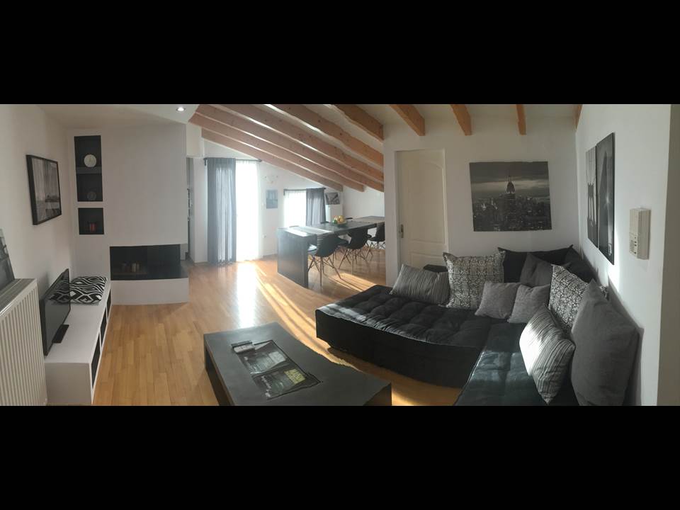 The spacious living area features a corner sofa with multiple cushions and a sleek coffee table. A fireplace adds warmth, while large windows allow natural light to fill the room. The dining area is visible in the background, designed for group meals.